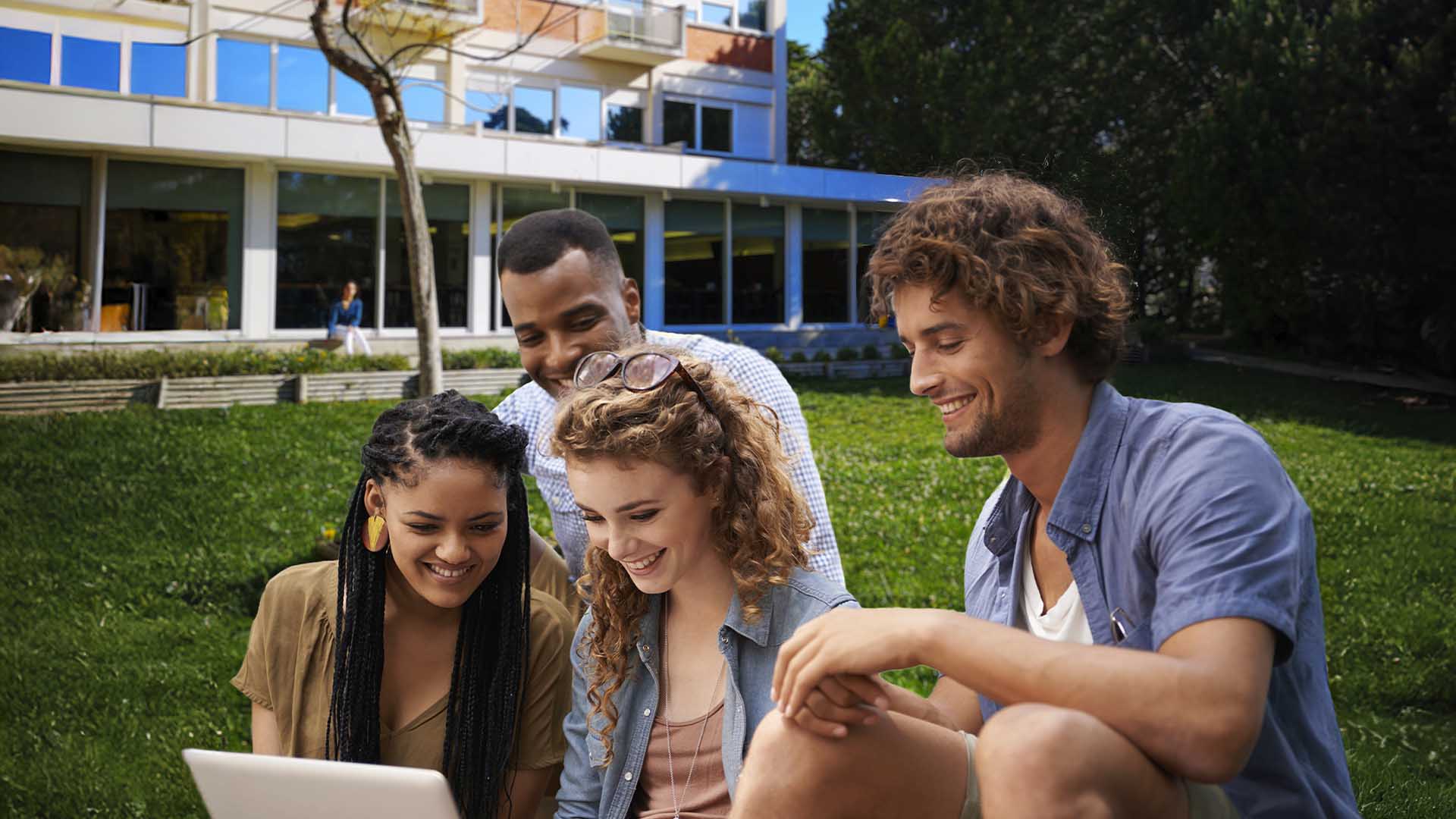 Quatro jovens sentados na relva, observando atentamente um laptop, em um ambiente ao ar livre e descontraído.