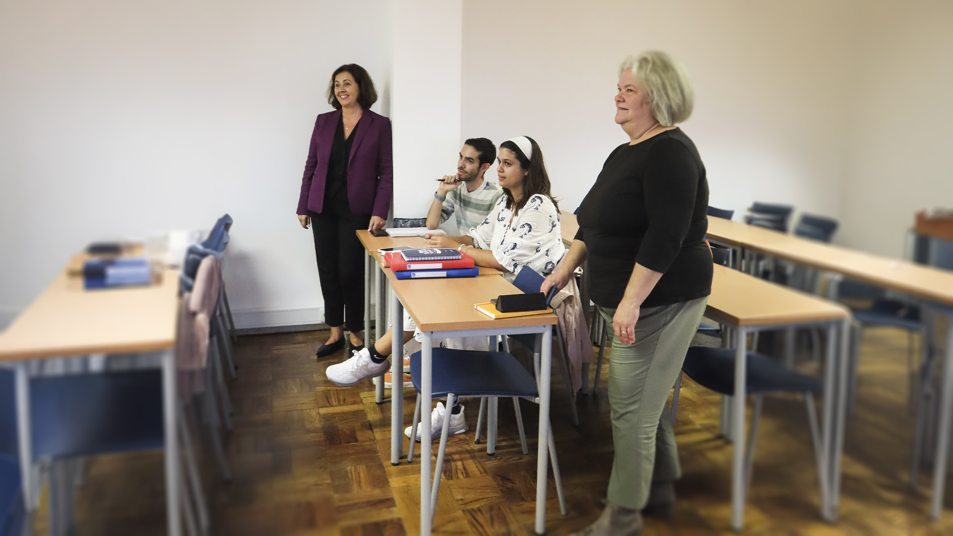 pessoas numa sala de aula, com duas mulheres em pé e dois alunos sentados à mesa.