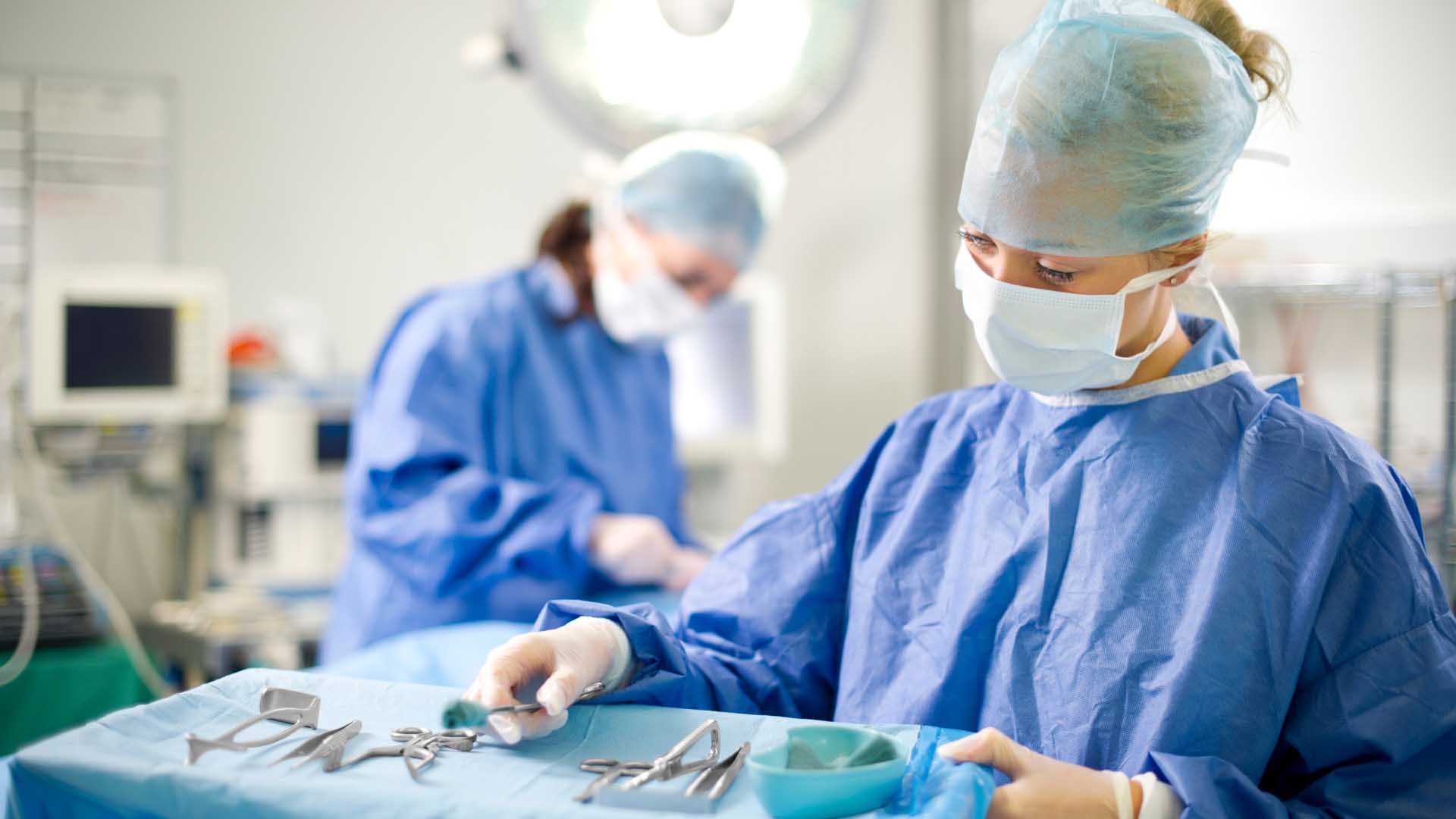 Profissionais de saúde com vestuário cirúrgico completo preparam instrumentos médicos numa mesa coberta por um pano azul, dentro de um bloco operatório iluminado.